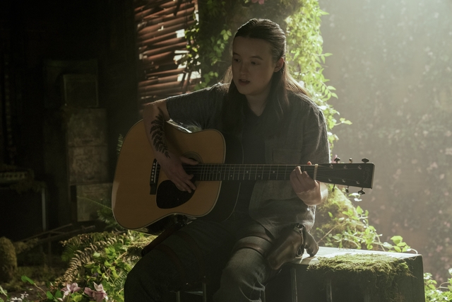 Ellie plays the guitar in a ruined building with light shining on her.