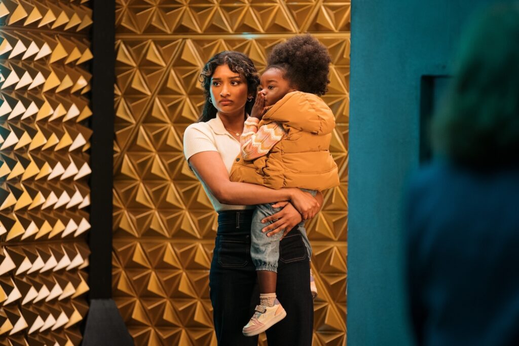 Belinda Chandra (Varada Sethu) holds Poppy (Sienna-Robyn Mavanga-Phipps) as she is about to step into a box that is meant to protect them from the events of The Reality War. Poppy blows a kiss to The Doctor, who is off screen.